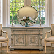 Load image into Gallery viewer, An aged silver round convex mirror on a wooden sideboard in front of a large bay window
