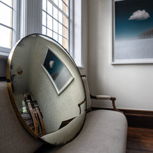 Load image into Gallery viewer, An aged silver mirror propped on a sofa and reflecting a painting  of blue sky and clouds
