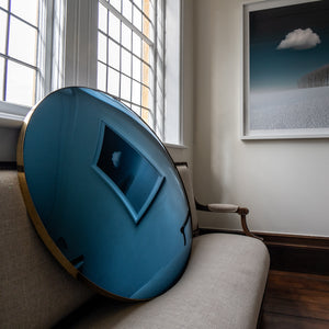 A Blue Round Convex Mirror propped on a traditional sofa. The Mirror reflects a landscape painting of  blue sky, trees and white clouds.