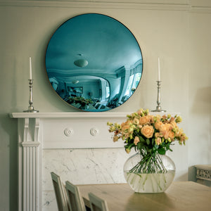 A Blue Round Convex Mirror placed on the mantelpiece of a traditional fireplace, and flanked by candles. A dining room table with a vase of yellow flowers is in the foreground.