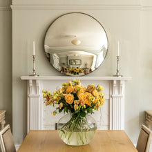 Load image into Gallery viewer, A Silver Round Convex Mirror propped on the  mantelpiece of a traditional fireplace with a dining table and vase of yellow flowers in the foreground