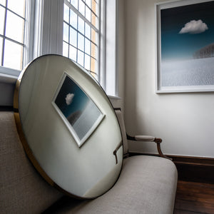 A Silver Round Convex Mirror propped on a traditional sofa and reflecting a wall landscape painting of blue sky and clouds