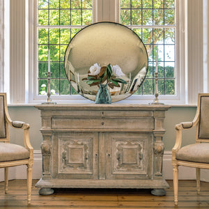 An aged silver round convex mirror on a wooden sideboard in front of a large bay window