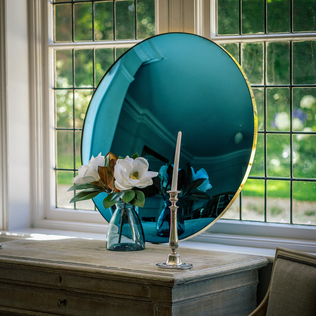 A Blue Round Convex Mirror propped on a wooden sideboard and against a large bay window. A vase of flowers and candle stick is placed in front of the mirror.