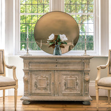 Load image into Gallery viewer, A Bronze Round Convex Mirror is propped on a rustic wooden sideboard and against a large bay window. A vase with two white flowers is on the sideboard  in front of the mirror and flanked by candles.
