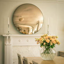 Load image into Gallery viewer, A Bronze Round Convex Mirror propped in a mantelpiece of a traditional fireplace and flanked by candles. A dining table, chairs and vase of yellow flowers is in the foreground.
