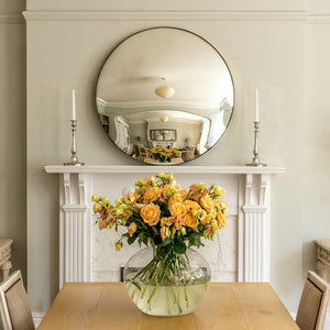 A Silver Round Convex Mirror propped on the  mantelpiece of a traditional fireplace with a dining table and vase of yellow flowers in the foreground